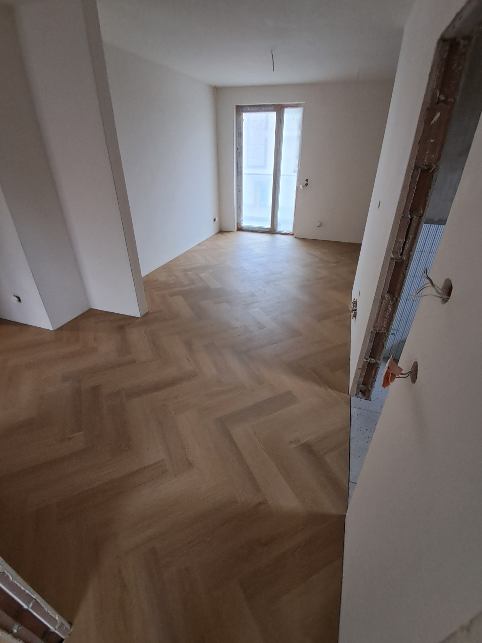 Jasny parkiet ułożony w jodełkę w pokoju z białymi ścianami i oknem. Widoczne fragmenty instalacji elektrycznej w ścianie. Ukończona podłoga w surowym wnętrzu.