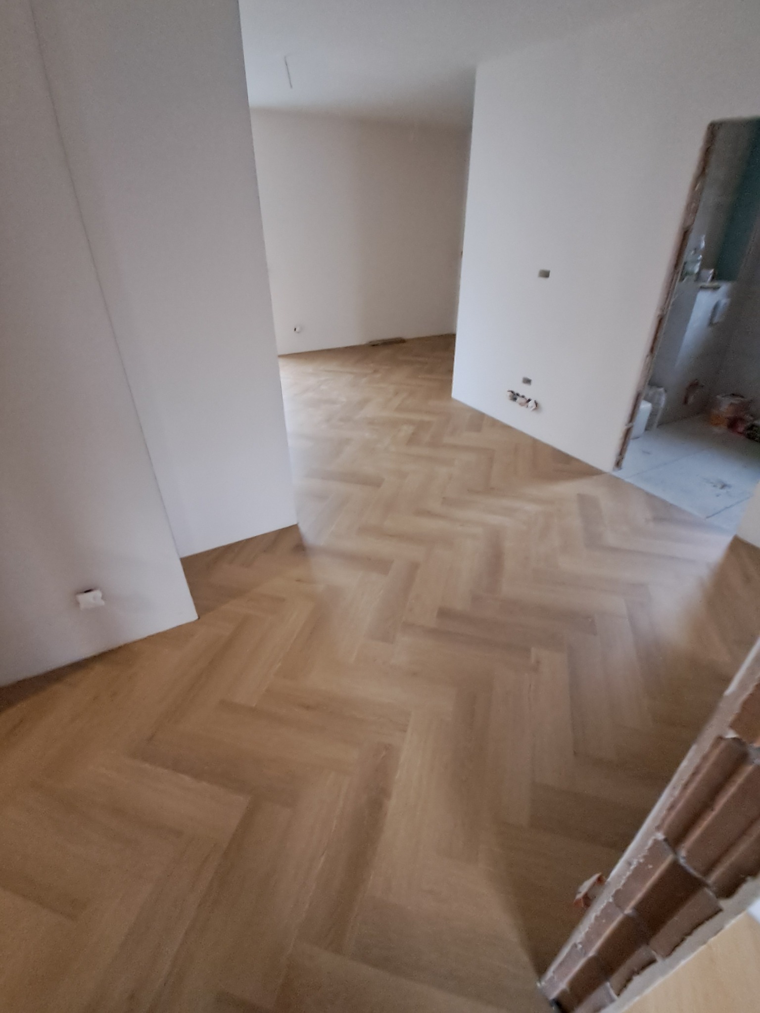 Jasny parkiet ułożony w jodełkę francuską w pustym, odnowionym pokoju. Widoczne gniazdka elektryczne i fragment futryny drzwiowej. Neutralna kolorystyka.