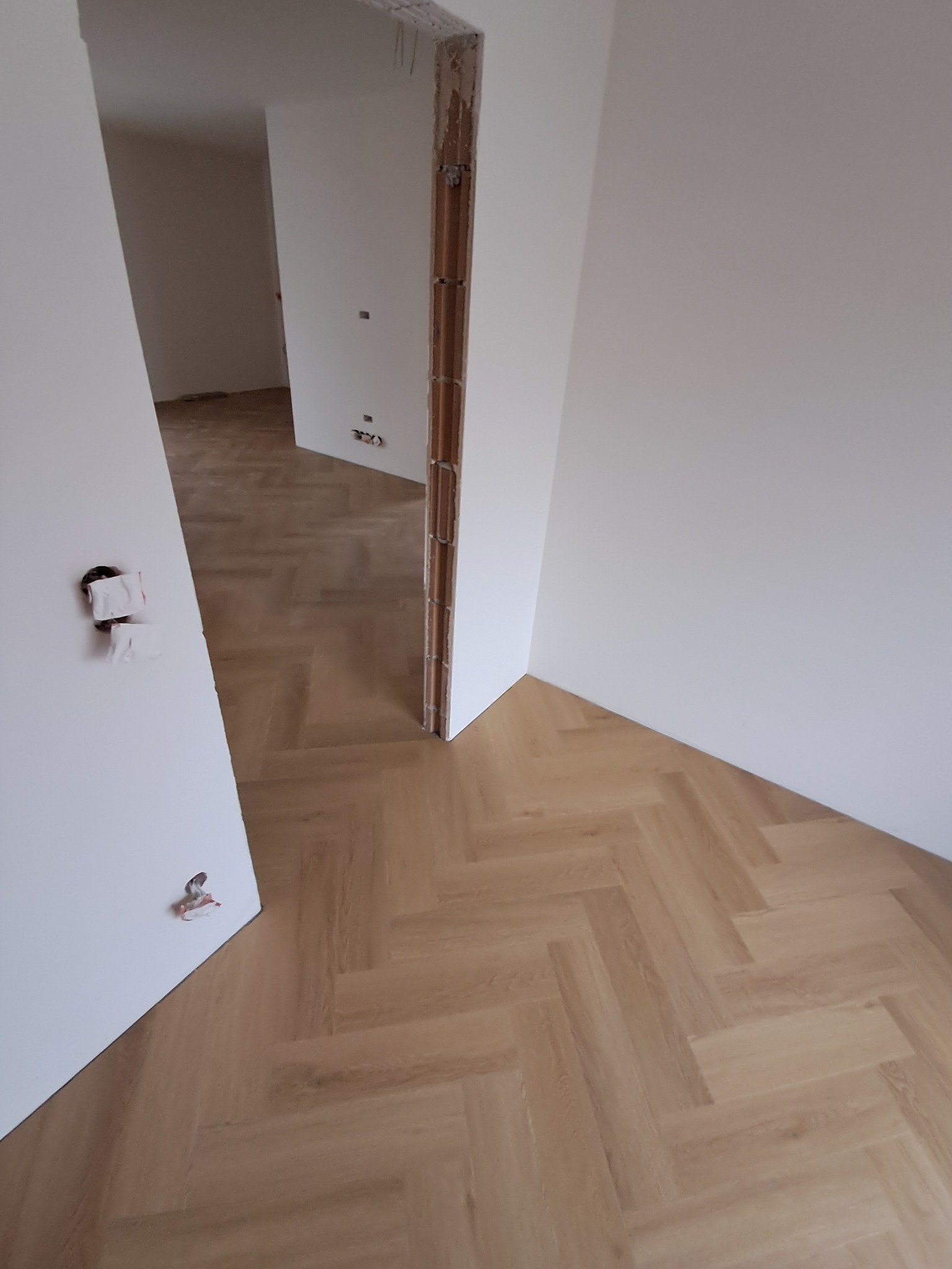 Jasny parkiet ułożony we wzór jodełki francuskiej w nowym wnętrzu. Widoczne surowe wykończenie ościeżnicy i wystające kable elektryczne.