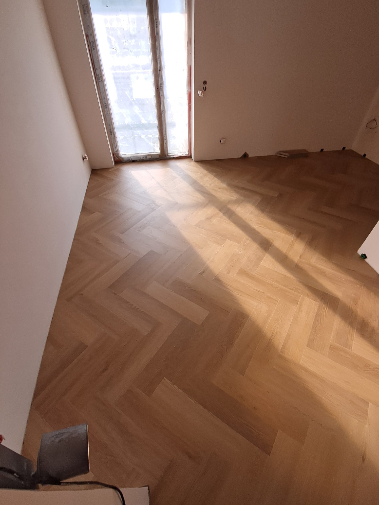 Ukończona podłoga z parkietu ułożonego w jodełkę w jasnym pokoju z oknem. Widoczne promienie słoneczne. Fragment narzędzia w lewym dolnym rogu.