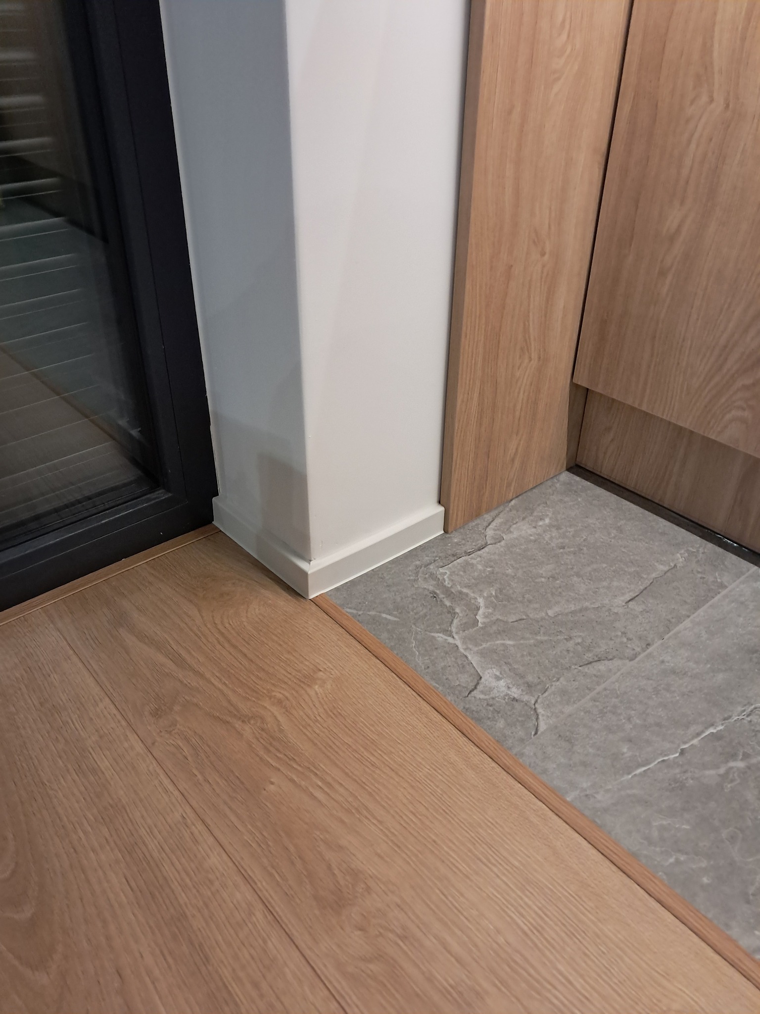 Połączenie paneli podłogowych z szarą, kamienną posadzką przy białej ścianie i czarnej ramie okiennej. Widoczne listwy przypodłogowe i wykończenie drewniane.