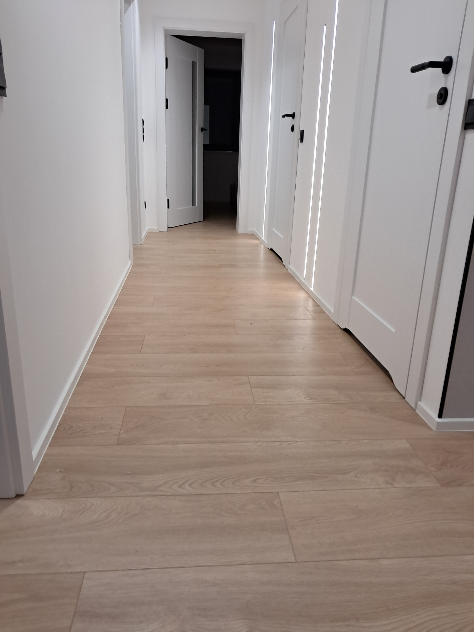 Jasne panele podłogowe w korytarzu z białymi ścianami i drzwiami z czarnymi klamkami. Widok z dołu podkreśla długość i perspektywę korytarza.