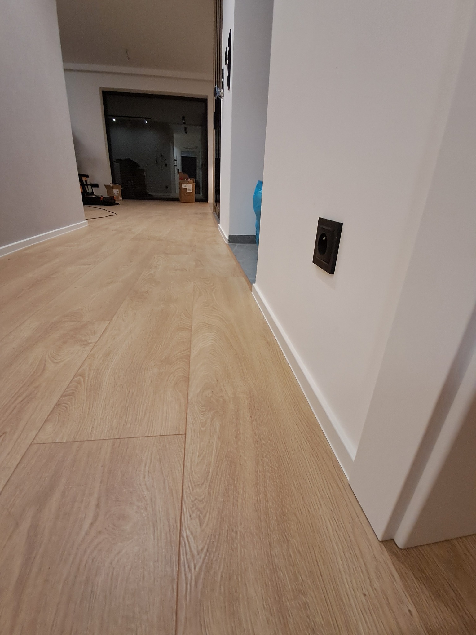 Beżowe panele podłogowe w korytarzu, widoczne listwy przypodłogowe i czarne gniazdko elektryczne na białej ścianie. W tle widoczne drzwi.
