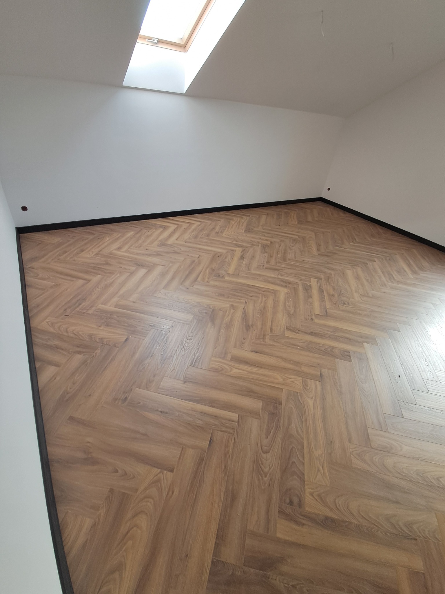 Ukończona podłoga z paneli ułożonych w jodełkę, wykończona czarnymi listwami przypodłogowymi w jasnym pokoju z oknem dachowym. Widok z góry.