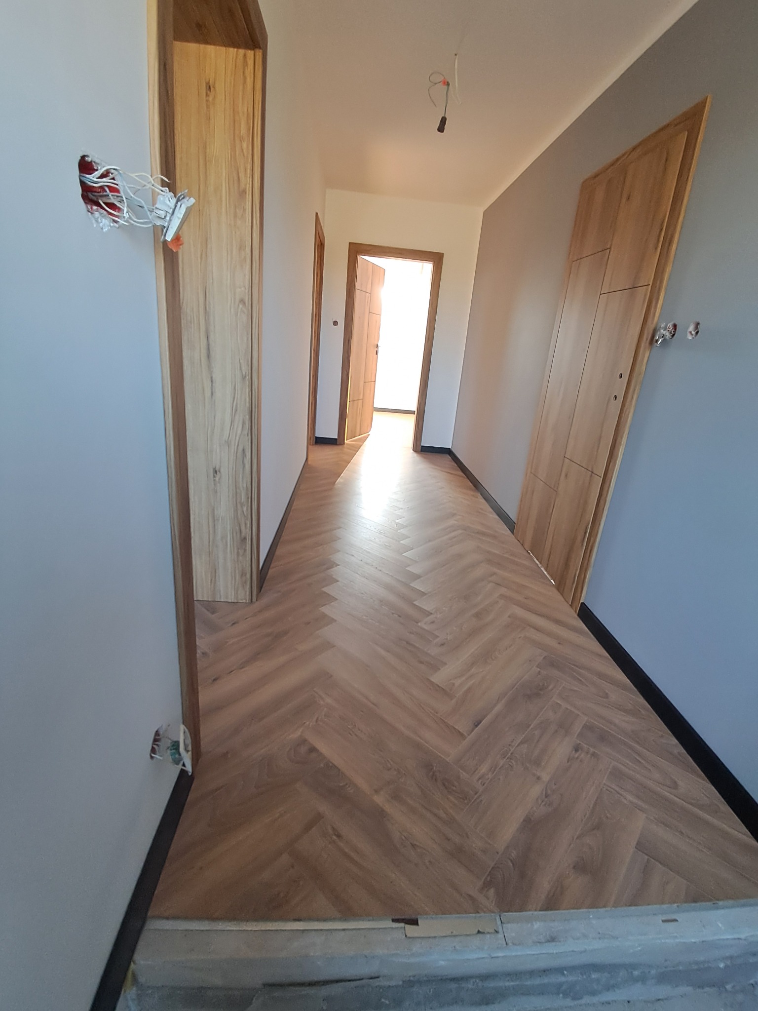 Korytarz po remoncie z parkietem ułożonym w jodełkę. Widoczne niedokończone prace elektryczne i futryny drzwi. Ściany pomalowane na szaro.