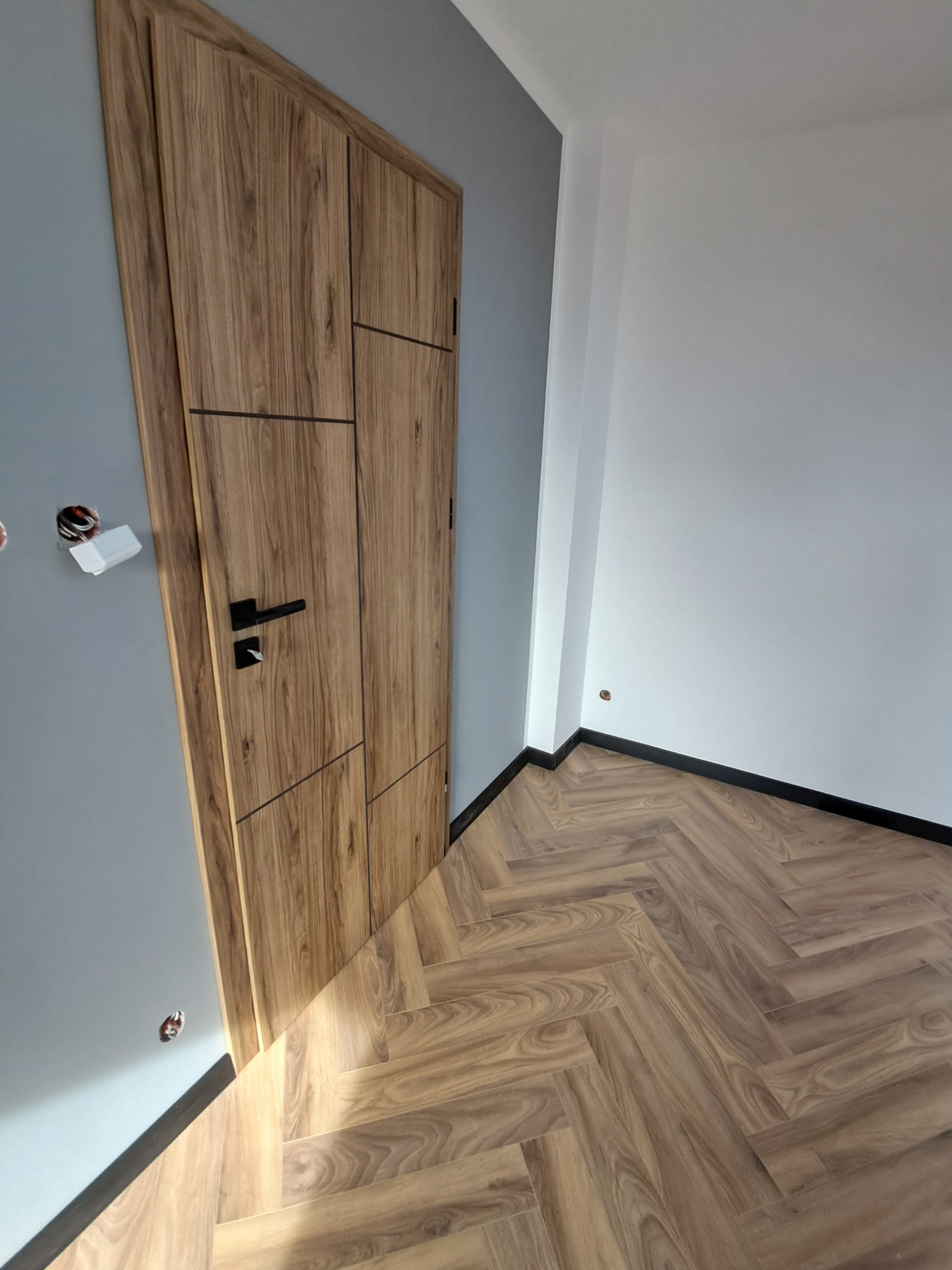 Nowoczesne wnętrze: drewniana podłoga ułożona w jodełkę, designerskie drzwi z drewnianym fornirem, szara ściana i białe listwy przypodłogowe.