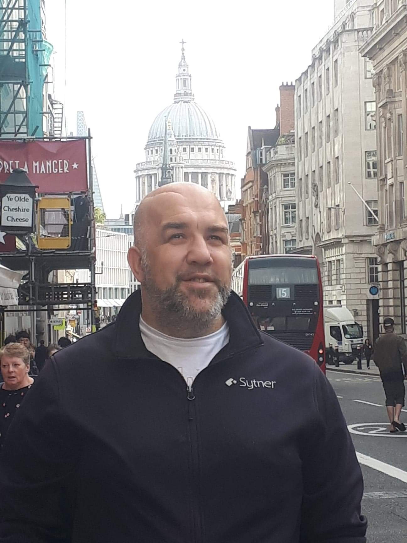 Mężczyzna w czarnej bluzie z logo 'Sytner' na tle londyńskiej ulicy z czerwonym autobusem i kopułą katedry św. Pawła w oddali.