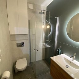 DOM 360 Damian Dobroć - Nowoczesna łazienka w szarościach z kabiną prysznicową typu walk-in, podwieszaną szafką i okrągłym lustrem z podświetleniem LED. Umywalka nablatowa z drewnianą szafką.