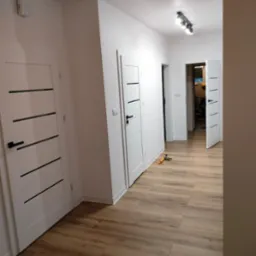 Jasne panele podłogowe w korytarzu po remoncie, białe ściany i nowoczesne drzwi z czarnymi akcentami, widoczne narzędzia glazurnicze.