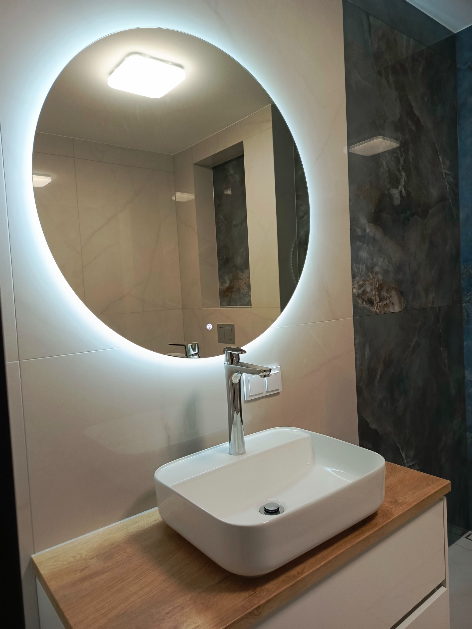 Nowoczesna łazienka: biała umywalka nablatowa, bateria i okrągłe lustro z podświetleniem LED na tle płytek imitujących marmur w odcieniach szarości.