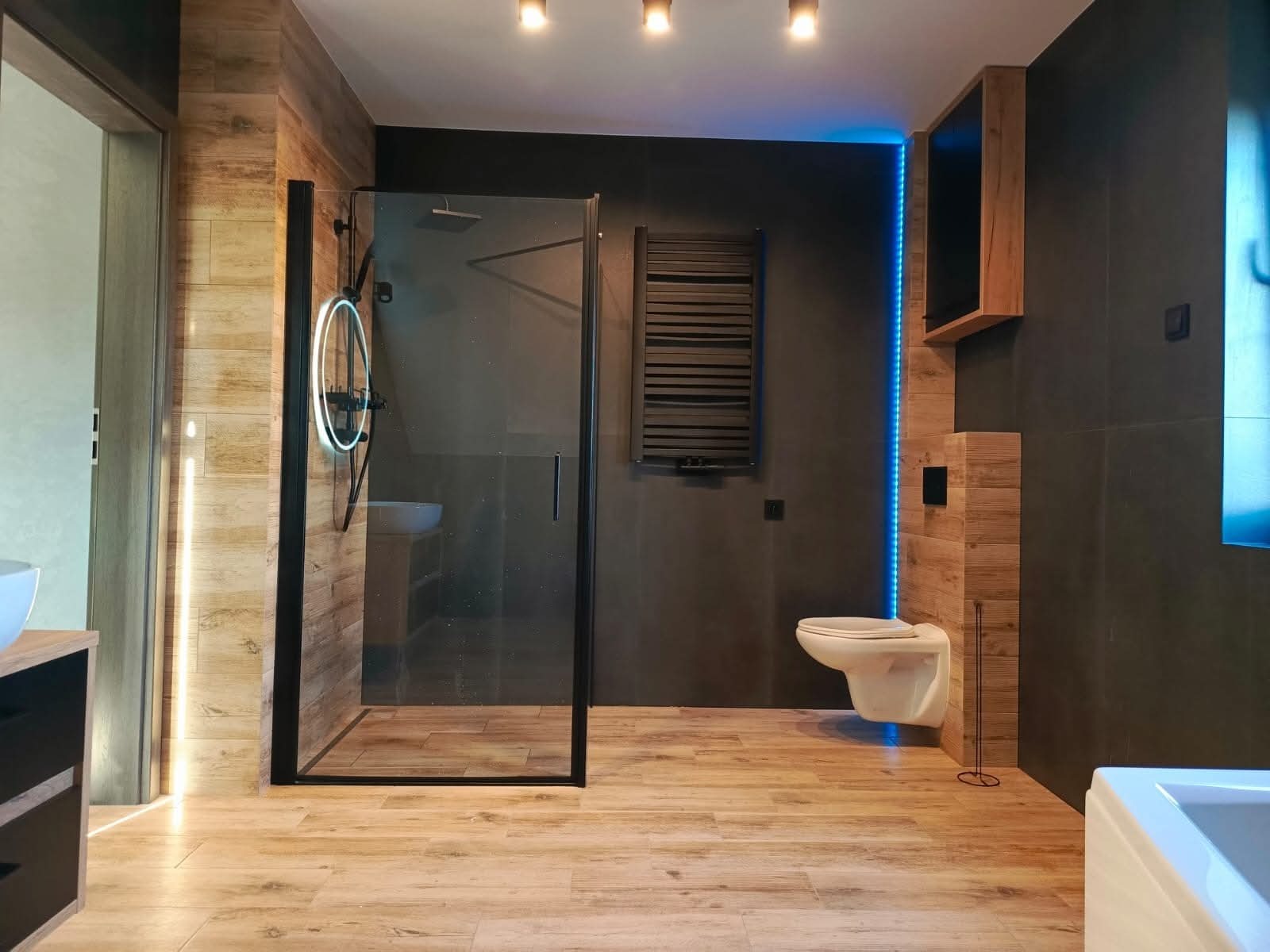 Nowoczesna łazienka z kabiną prysznicową typu walk-in, drewnopodobnymi płytkami i czarnymi akcentami, podświetlona listwą LED. Umywalka z okrągłym lustrem.