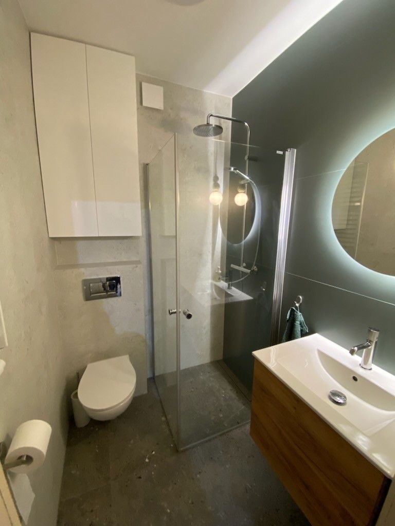 Nowoczesna łazienka w szarościach z kabiną prysznicową typu walk-in, podwieszaną szafką i okrągłym lustrem z podświetleniem LED. Umywalka nablatowa z drewnianą szafką.