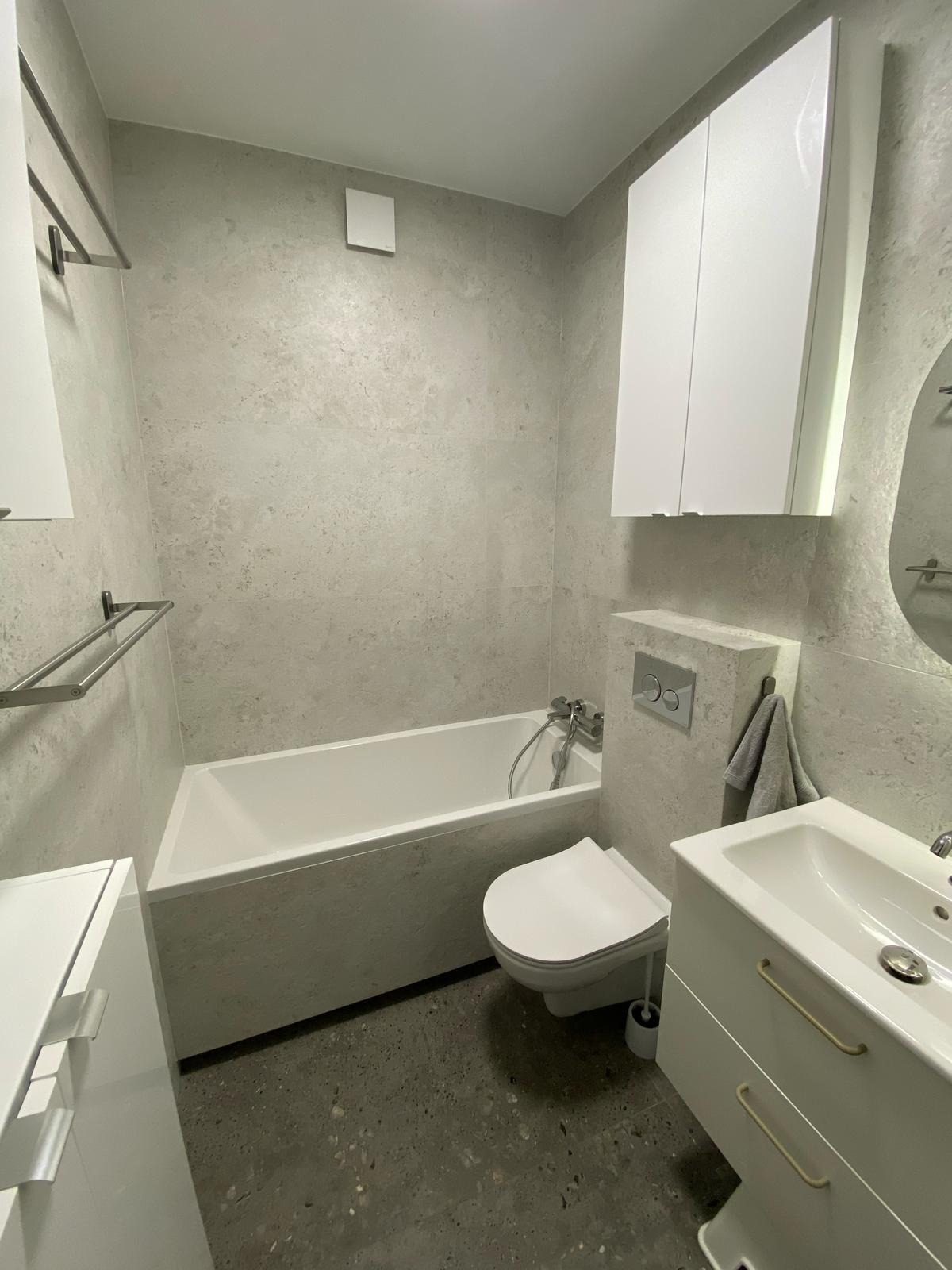 Nowoczesna łazienka w odcieniach szarości z białą armaturą i płytkami imitującymi kamień. Minimalistyczny design, wanna zabudowana, umywalka z szafką i wiszące szafki.