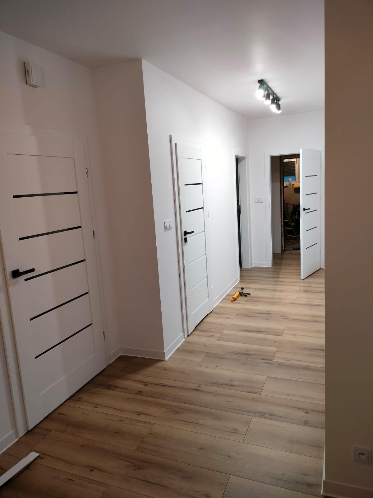 Jasne panele podłogowe w korytarzu po remoncie, białe ściany i nowoczesne drzwi z czarnymi akcentami, widoczne narzędzia glazurnicze.