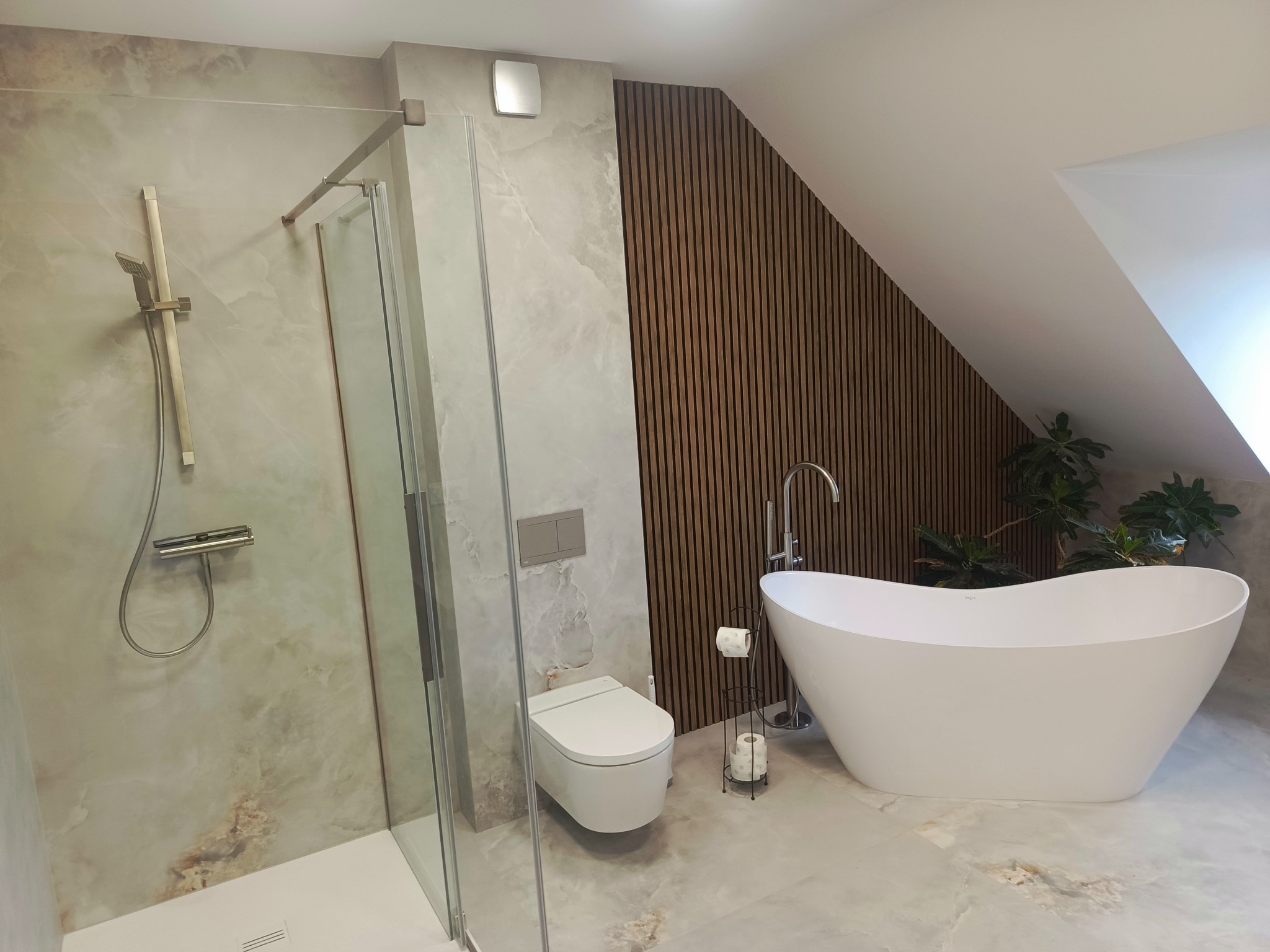 Elegancka łazienka na poddaszu z kabiną prysznicową, wolnostojącą wanną i drewnianymi panelami ściennymi. Marmurowe płytki podłogowe i rośliny doniczkowe.