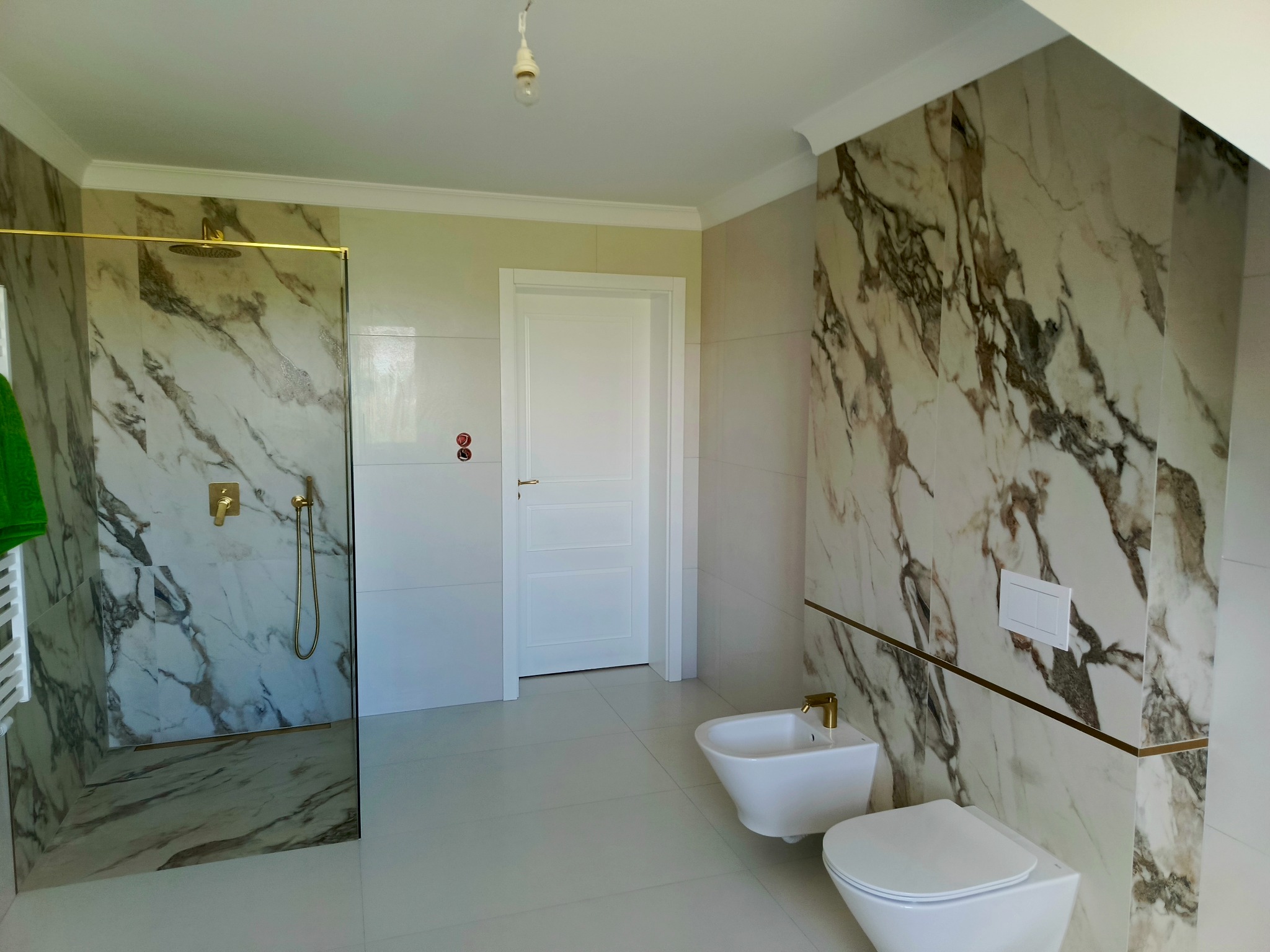 Elegancka łazienka z marmurowymi płytkami, złotymi akcentami armatury i kabiną prysznicową typu walk-in. Widoczne białe sanitariaty i minimalistyczny design.