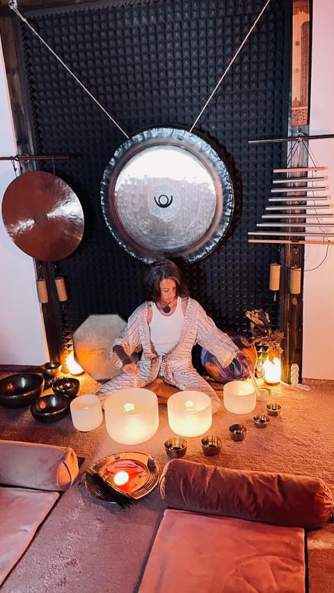 Kobieta siedząca na podłodze w otoczeniu gongów, mis kryształowych i innych instrumentów dźwiękowych podczas sesji relaksacyjnej.