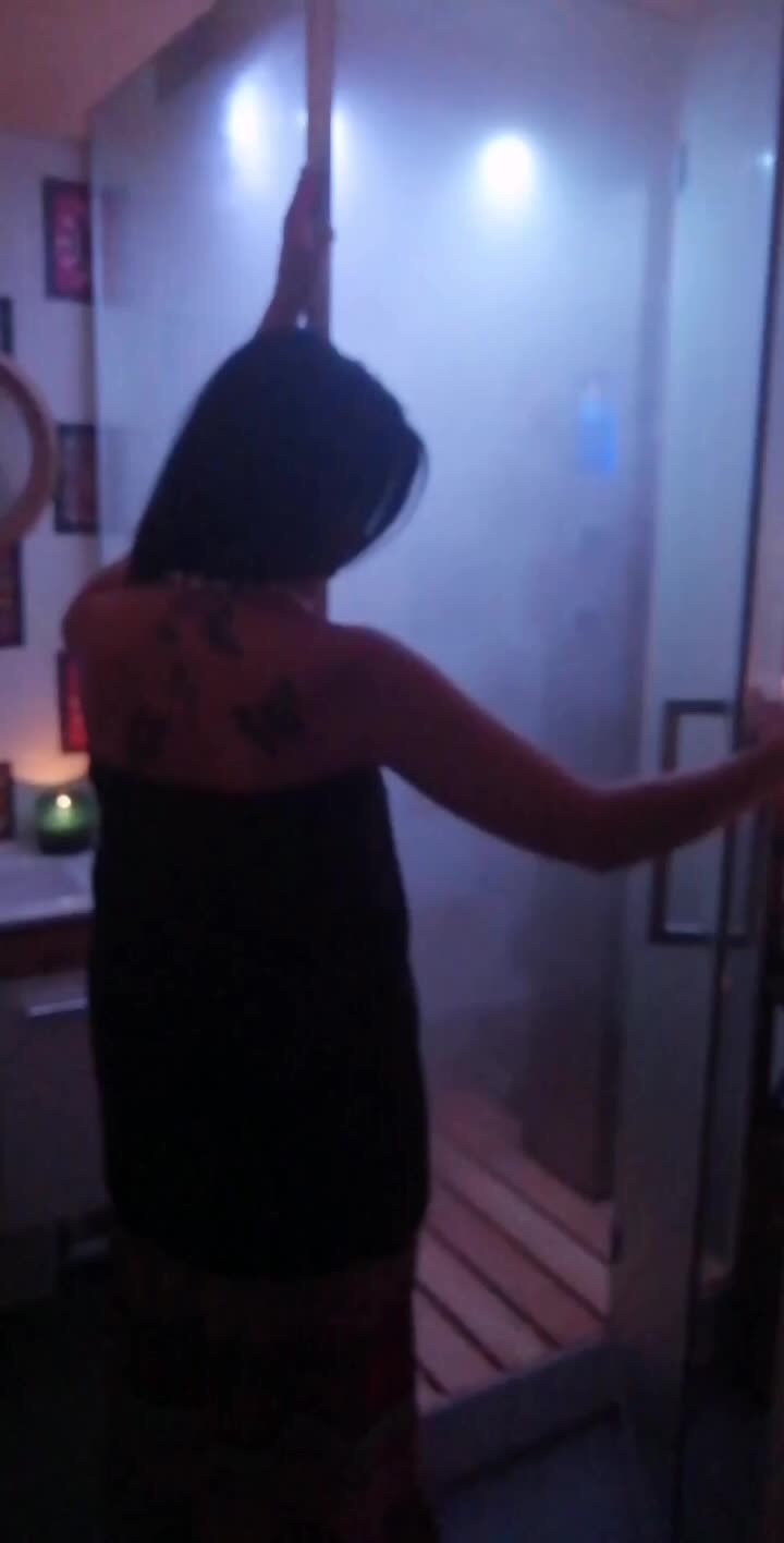 Kobieta owinięta ręcznikiem wchodzi do sauny parowej z drewnianymi deskami, widoczne tatuaże na plecach, wnętrze oświetlone niebieskim światłem.