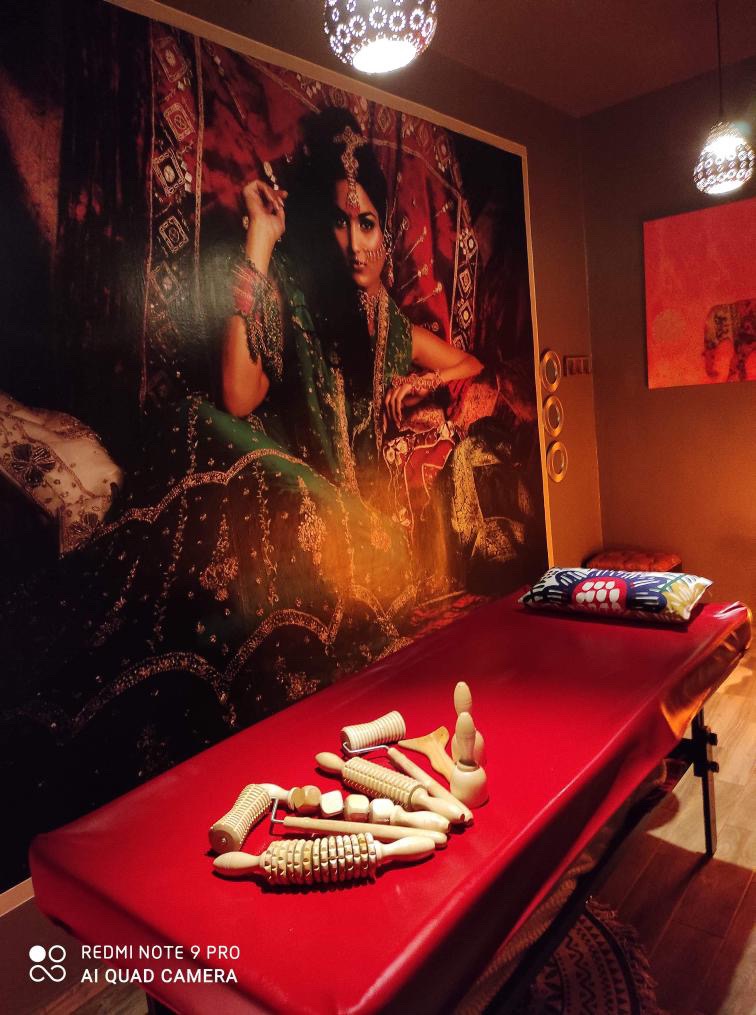 Czerwony stół do zabiegów z drewnianymi wałkami do masażu, orientalny wystrój wnętrza z dużym zdjęciem kobiety w tradycyjnym stroju indyjskim na ścianie.