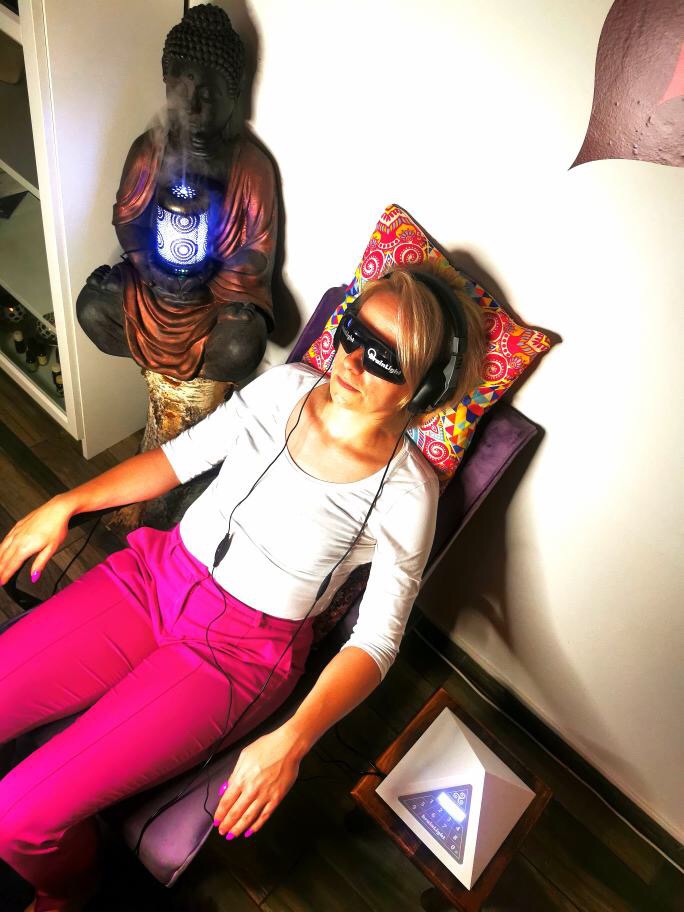 Kobieta w różowych spodniach relaksuje się na fotelu z maską na oczach i słuchawkami, obok stoi podświetlana statuetka Buddy z efektem dymu oraz piramida ze światłem.