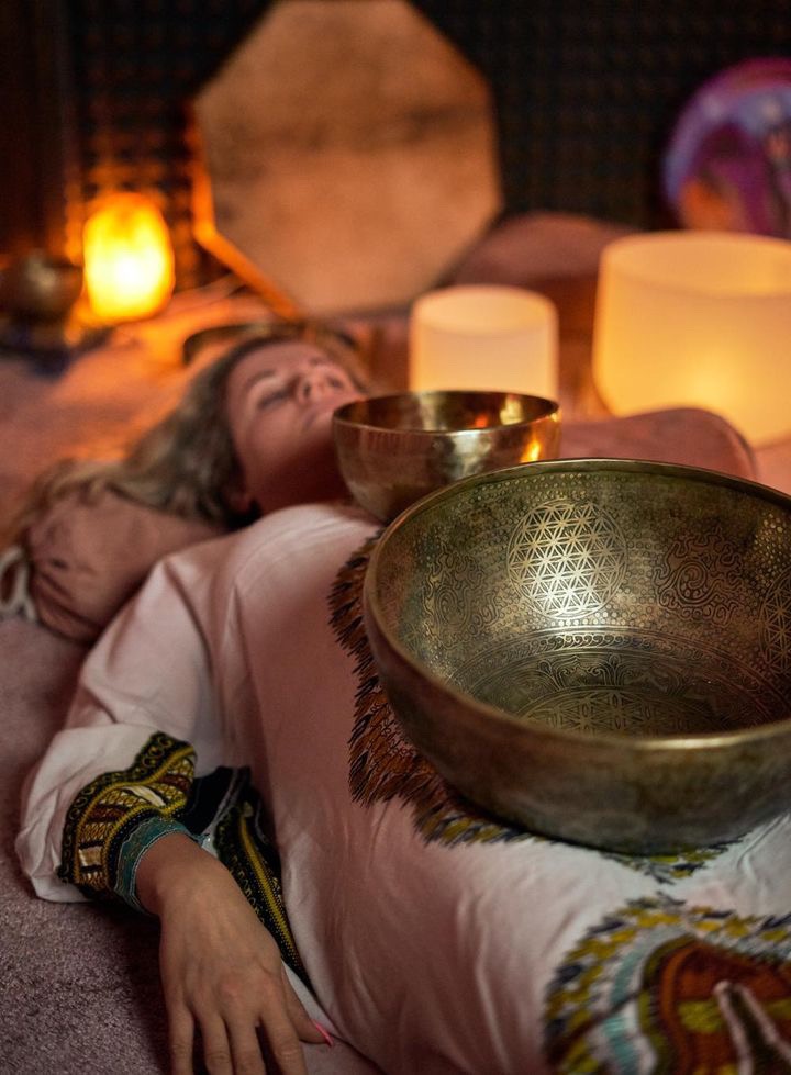 Kobieta leży na plecach z misami dźwiękowymi na ciele, w tle świece i instrumenty muzyczne, tworząc relaksującą atmosferę.
