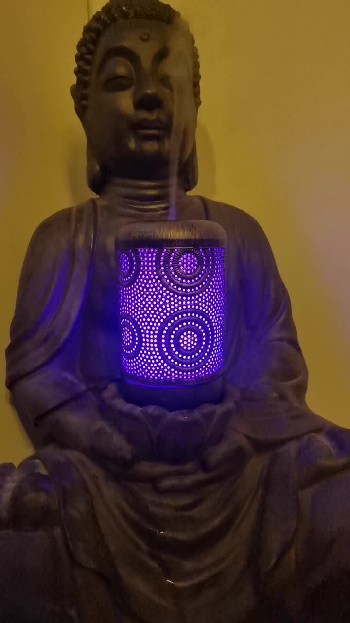 Statuetka Buddy z ciemnego kamienia, trzymająca w dłoniach zapalony, ażurowy dyfuzor z fioletowym światłem i wydobywającą się parą.