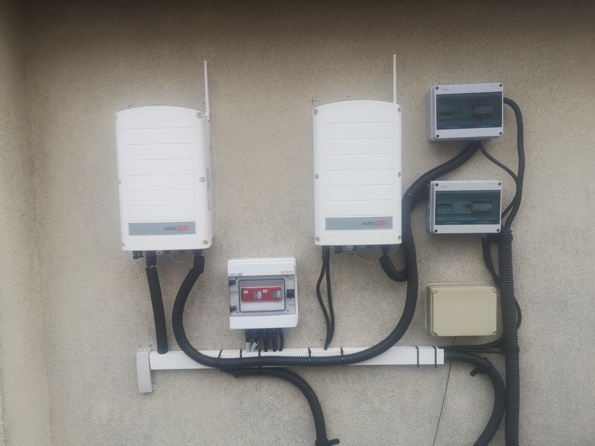 Dwa białe inwertery z antenami, zamontowane na zewnątrz budynku obok szarych skrzynek elektrycznych i peszli, przymocowanych do ściany za pomocą białej listwy.