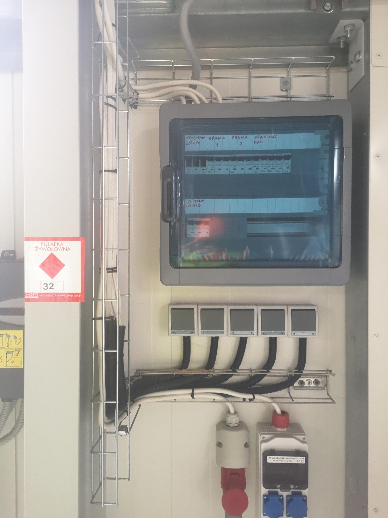 Rozdzielnia elektryczna z wyłącznikami oznaczonymi jako 'Wyłącznik główny', 'Brama 1', 'Brama 2', 'Oświetlenie hali', z zainstalowanymi gniazdami siłowymi i przewodami poprowadzonymi w korytkach...
