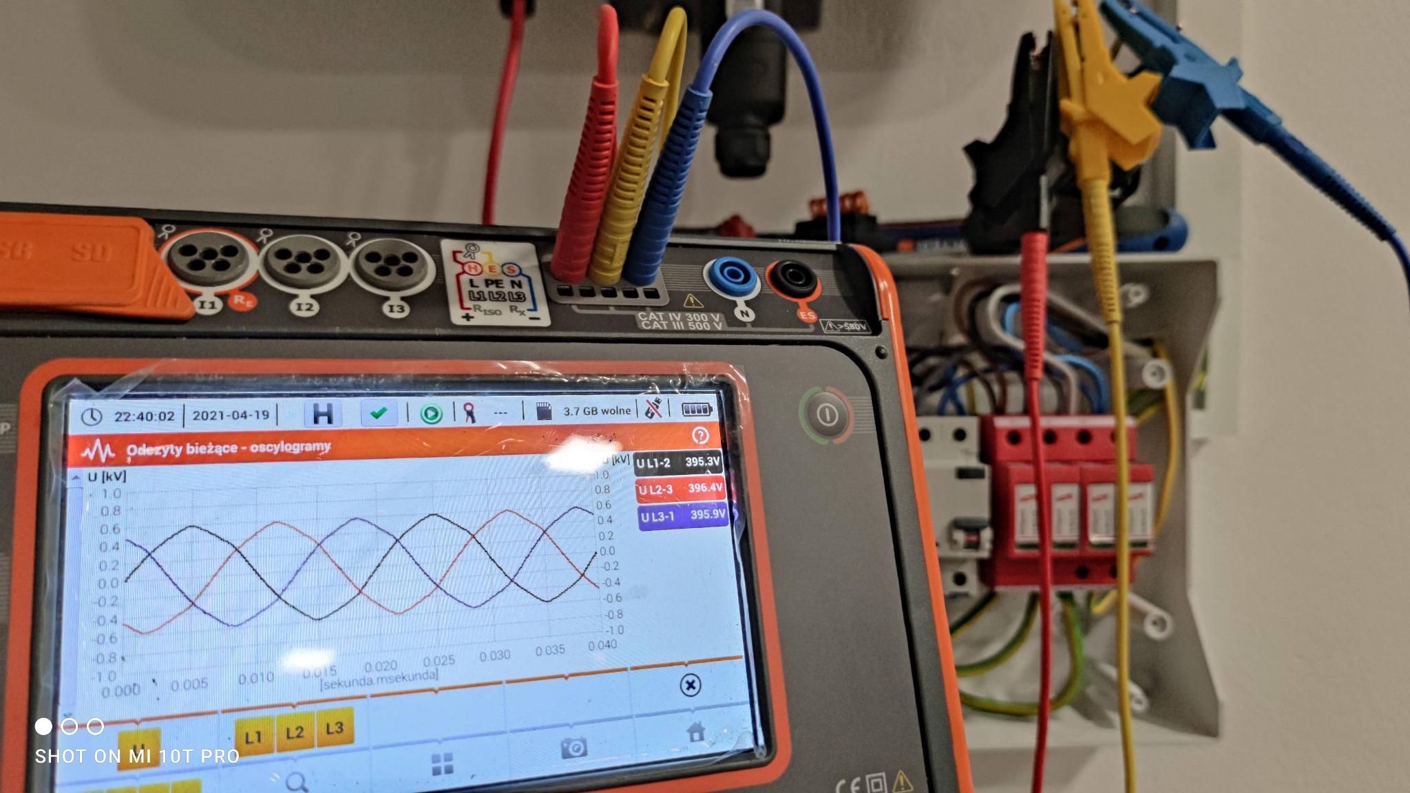 Miernik elektryczny z kolorowymi przewodami pomiarowymi podłączony do instalacji elektrycznej, wyświetlający oscylogramy napięć międzyfazowych.