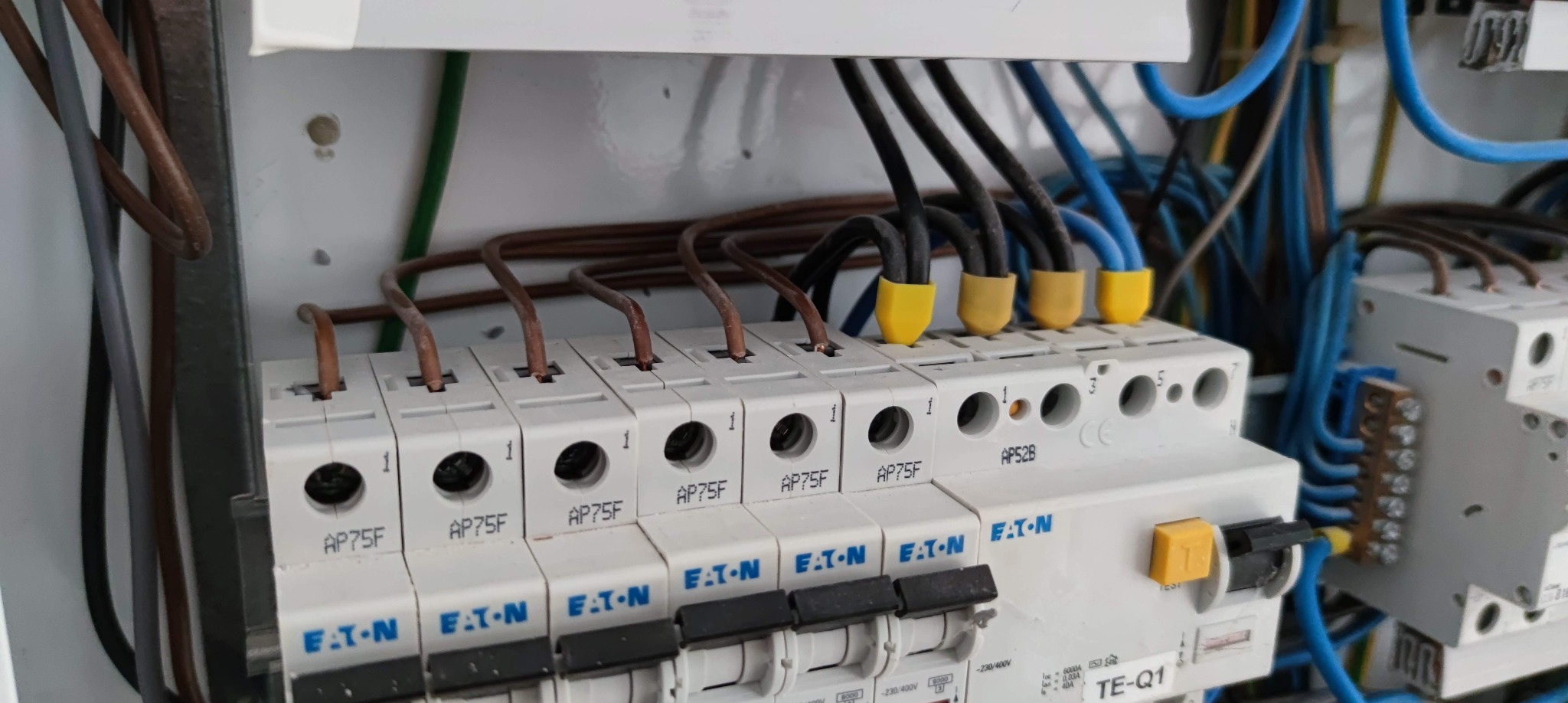 Szczegółowe ujęcie skrzynki elektrycznej z widocznymi bezpiecznikami EATON AP75F i AP52B, brązowymi przewodami oraz niebieskimi i czarnymi przewodami z żółtymi końcówkami, zamontowanymi w białej...