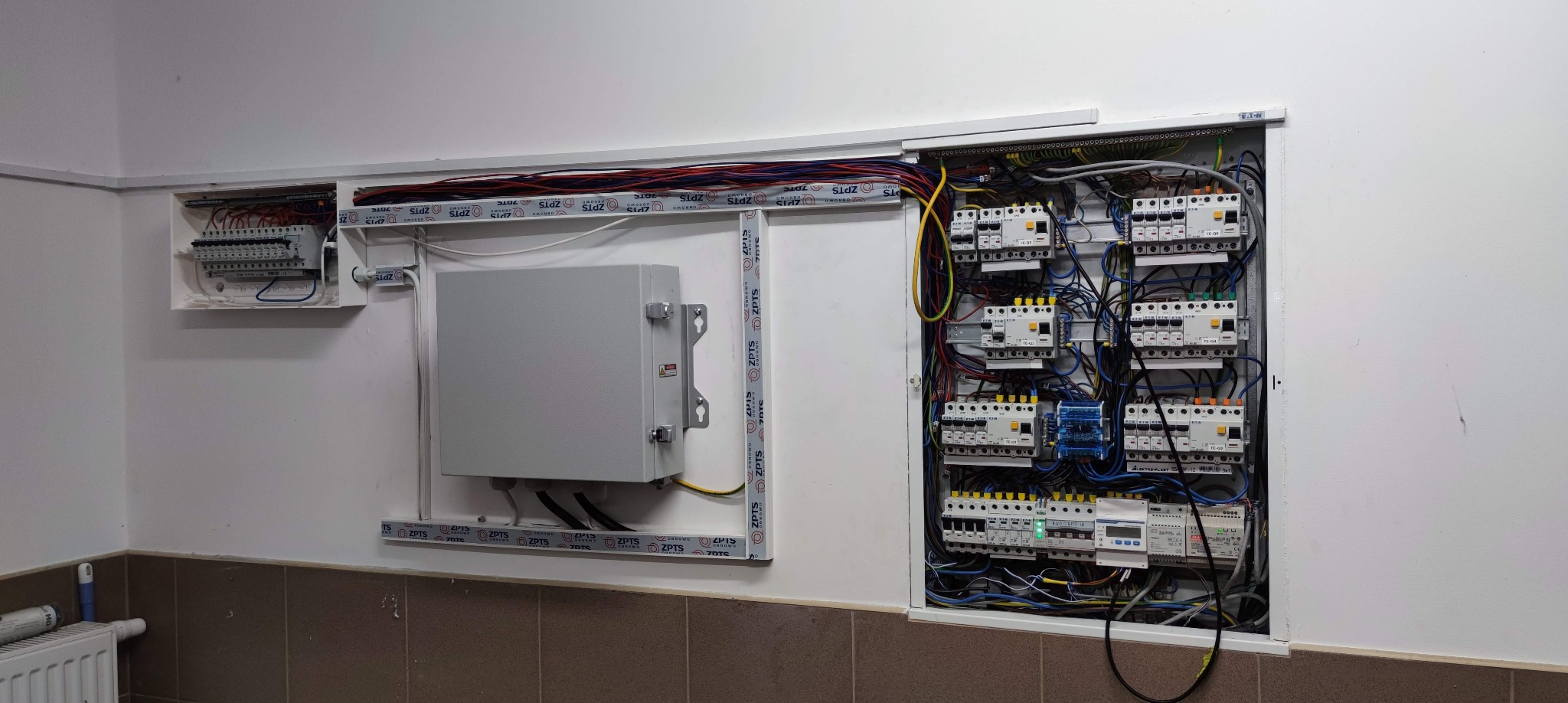 Rozdzielnica elektryczna z zabezpieczeniami i przewodami, umieszczona w ścianie, widoczne dodatkowe elementy i okablowanie w korytkach.