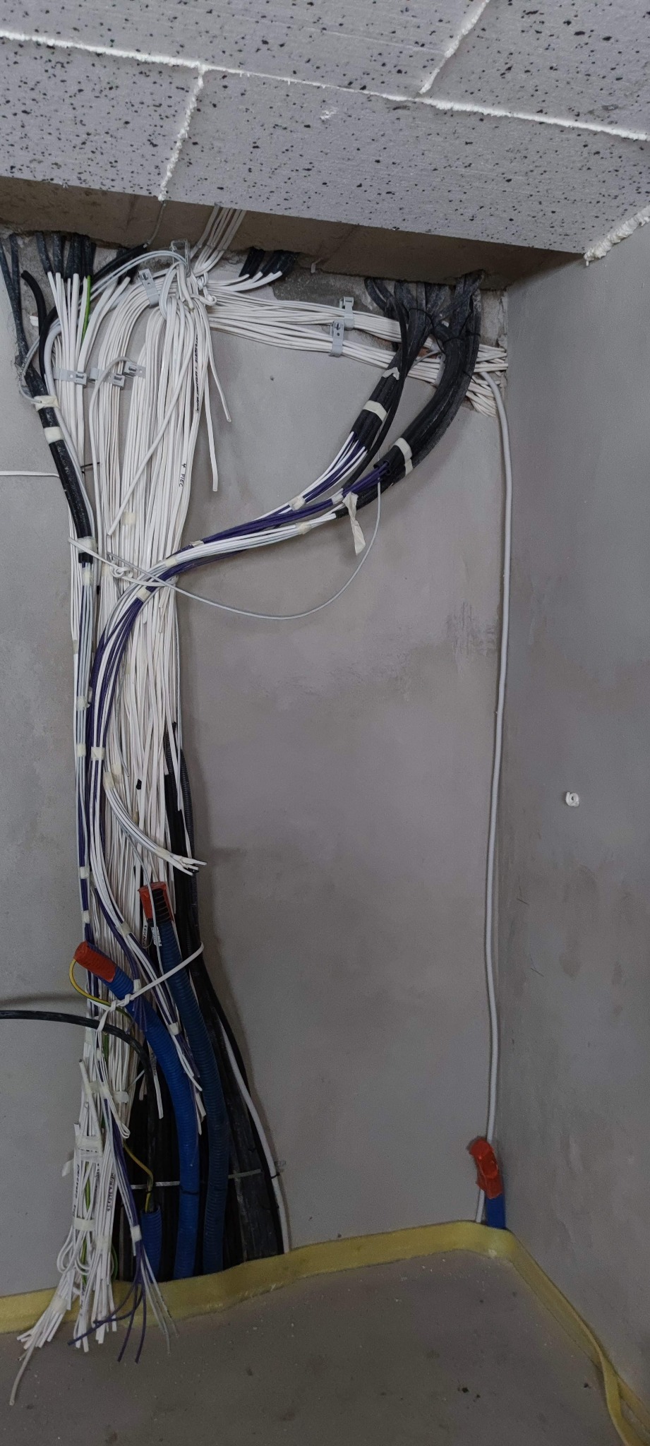 Wiązka kabli elektrycznych i rur instalacyjnych, prowadzonych wzdłuż szarej ściany, częściowo ukryta w suficie z płyt. Widoczne białe i czarne kable, niebieskie rury karbowane oraz pomarańczowe...