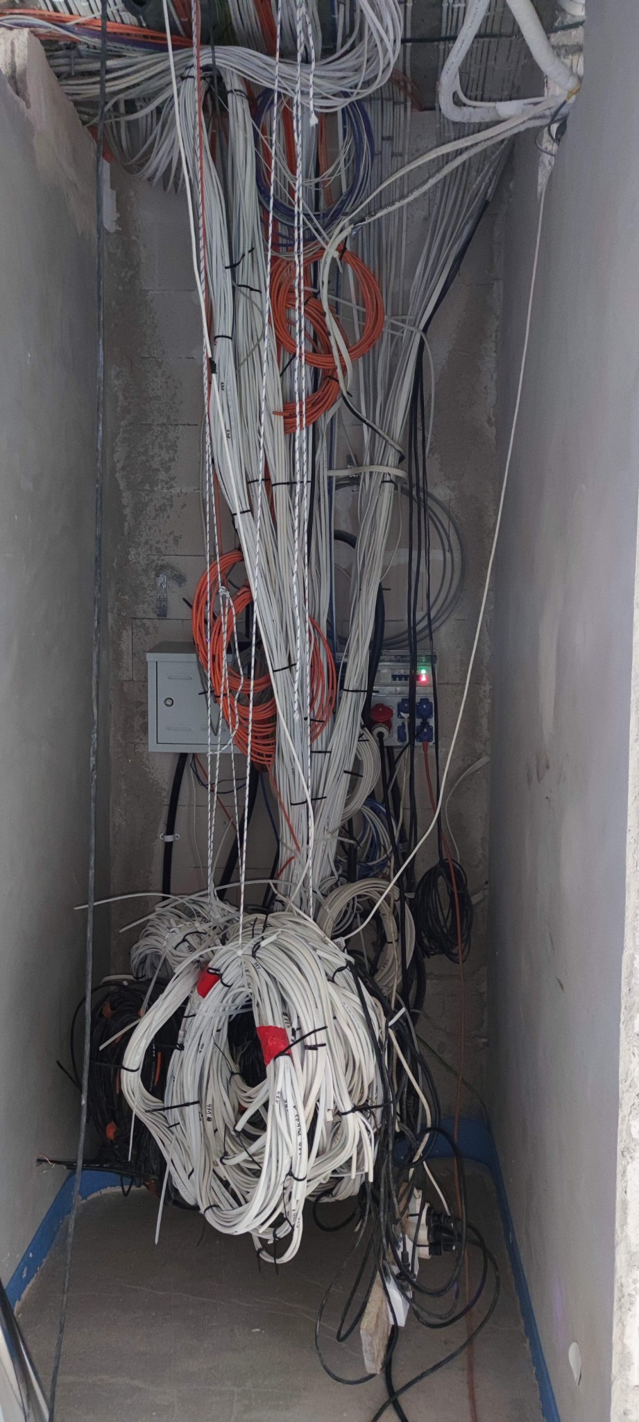Spiętrzenie przewodów elektrycznych w szarej wnęce ściany, widoczna skrzynka rozdzielcza z podłączonymi kablami, część przewodów zwinięta w pętle, część oznaczona czerwonymi taśmami.
