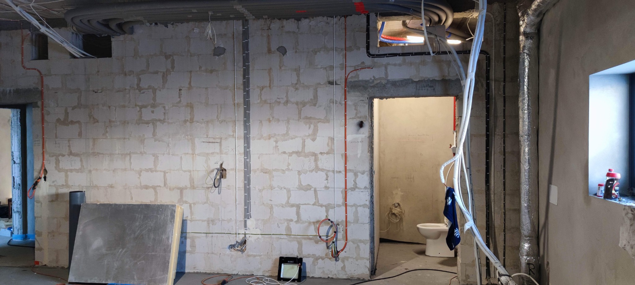 Surowy stan wnętrza w trakcie prac instalacyjnych: widoczne przewody elektryczne prowadzone po ścianach z bloczków, rury wentylacyjne w izolacji, przygotowane otwory w ścianach oraz niedokończona...