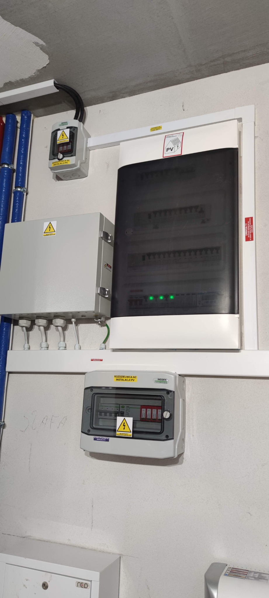 Rozdzielnice elektryczne AC i DC instalacji fotowoltaicznej, zamontowane na ścianie wewnątrz budynku, z widocznymi zabezpieczeniami i okablowaniem.
