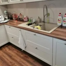 Fragment kuchni z białymi szafkami z frezowanymi frontami i drewnianym blatem, widoczny zlew z baterią, naczynia i płyny do mycia naczyń.