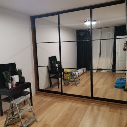Dan-Mar - Montaż dużej szafy przesuwnej z lustrami w czarnej ramie, widoczne narzędzia i akcesoria montażowe, podłoga wyłożona panelami.