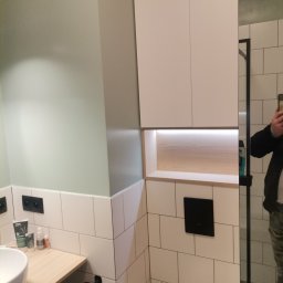 Dan-Mar - Nowoczesna łazienka z białymi, prostokątnymi płytkami, podwieszaną szafką z oświetleniem LED, umywalką nablatową i toaletą podwieszaną z czarnym przyciskiem spłukującym.