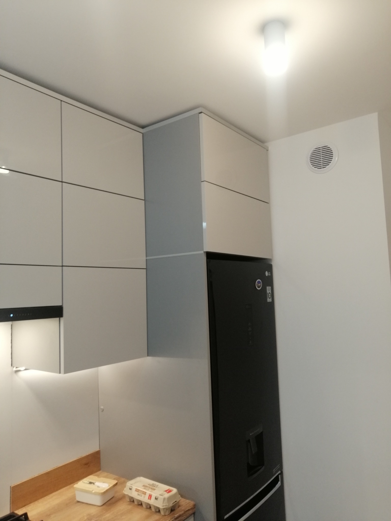 Nowoczesna kuchnia z białymi, lakierowanymi szafkami wiszącymi, czarną lodówką LG i drewnianym blatem z pojemnikami na żywność.