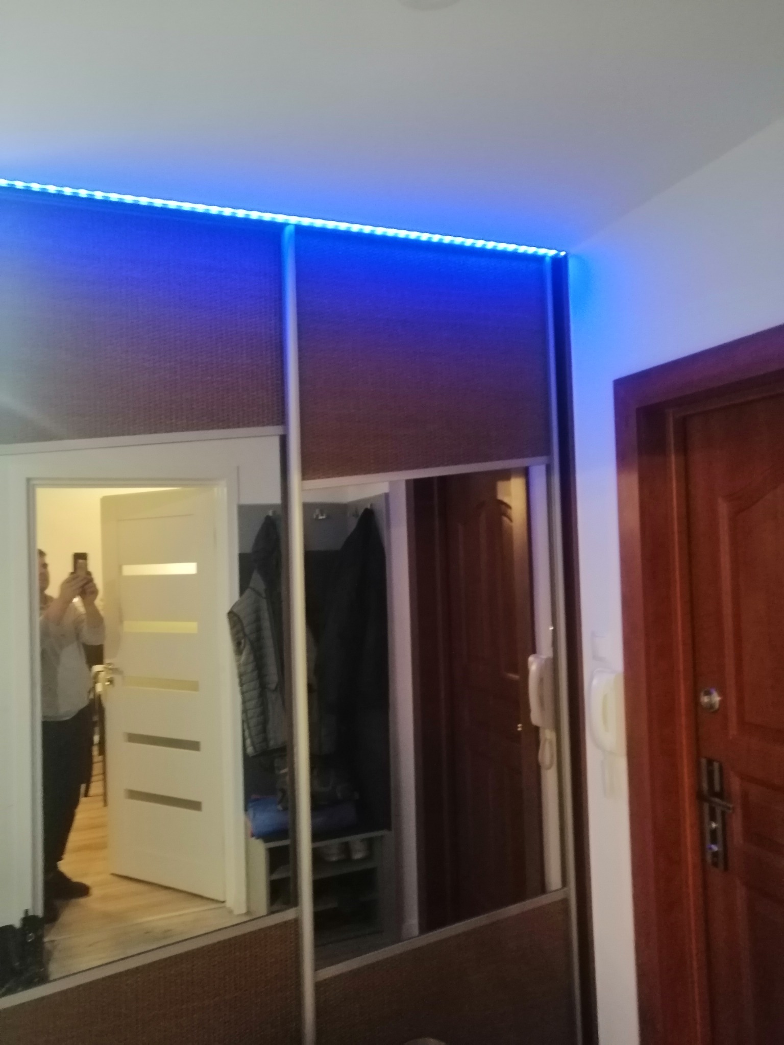 Wnętrze przedpokoju z szafą wnękową z przesuwnymi drzwiami lustrzanymi i oświetleniem LED, odbicie osoby robiącej zdjęcie telefonem.