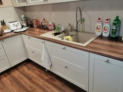Fragment kuchni z białymi szafkami z frezowanymi frontami i drewnianym blatem, widoczny zlew z baterią, naczynia i płyny do mycia naczyń.