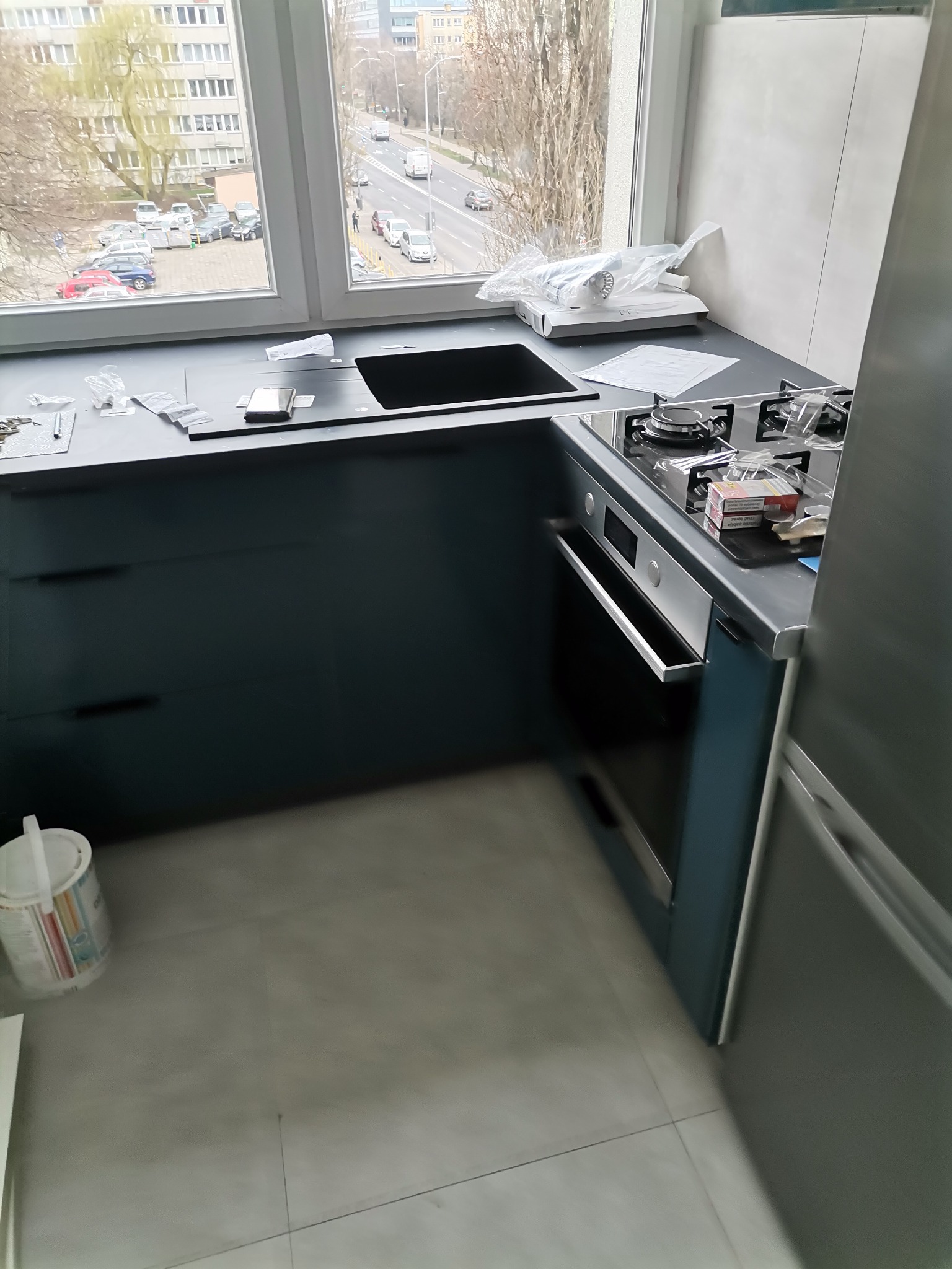 Montaż nowej, ciemnogranatowej zabudowy kuchennej z czarnym zlewozmywakiem i płytą gazową, widok z okna na miasto, biała puszka z farbą na podłodze.
