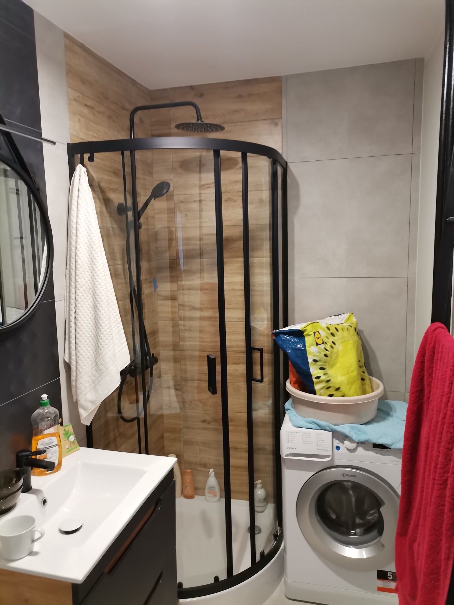 Nowoczesna łazienka po remoncie z kabiną prysznicową typu walk-in z czarnymi profilami, umywalką z szafką w kolorze antracytowym i pralką Indesit.