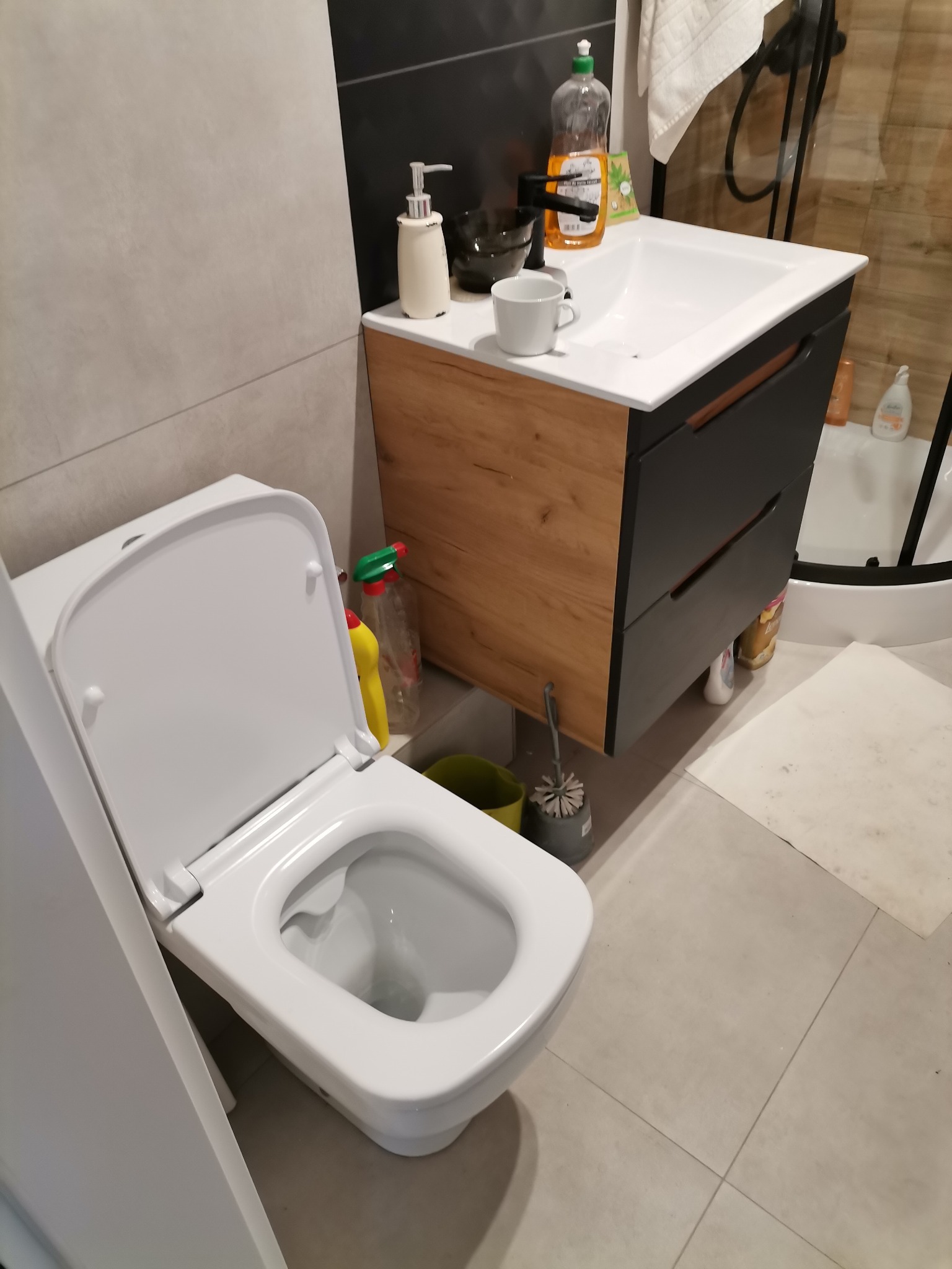 Nowoczesna łazienka z podwieszaną toaletą z otwartą deską, umywalką nablatową na szafce z drewnopodobnym frontem i ciemnymi szufladami oraz szare płytki podłogowe.