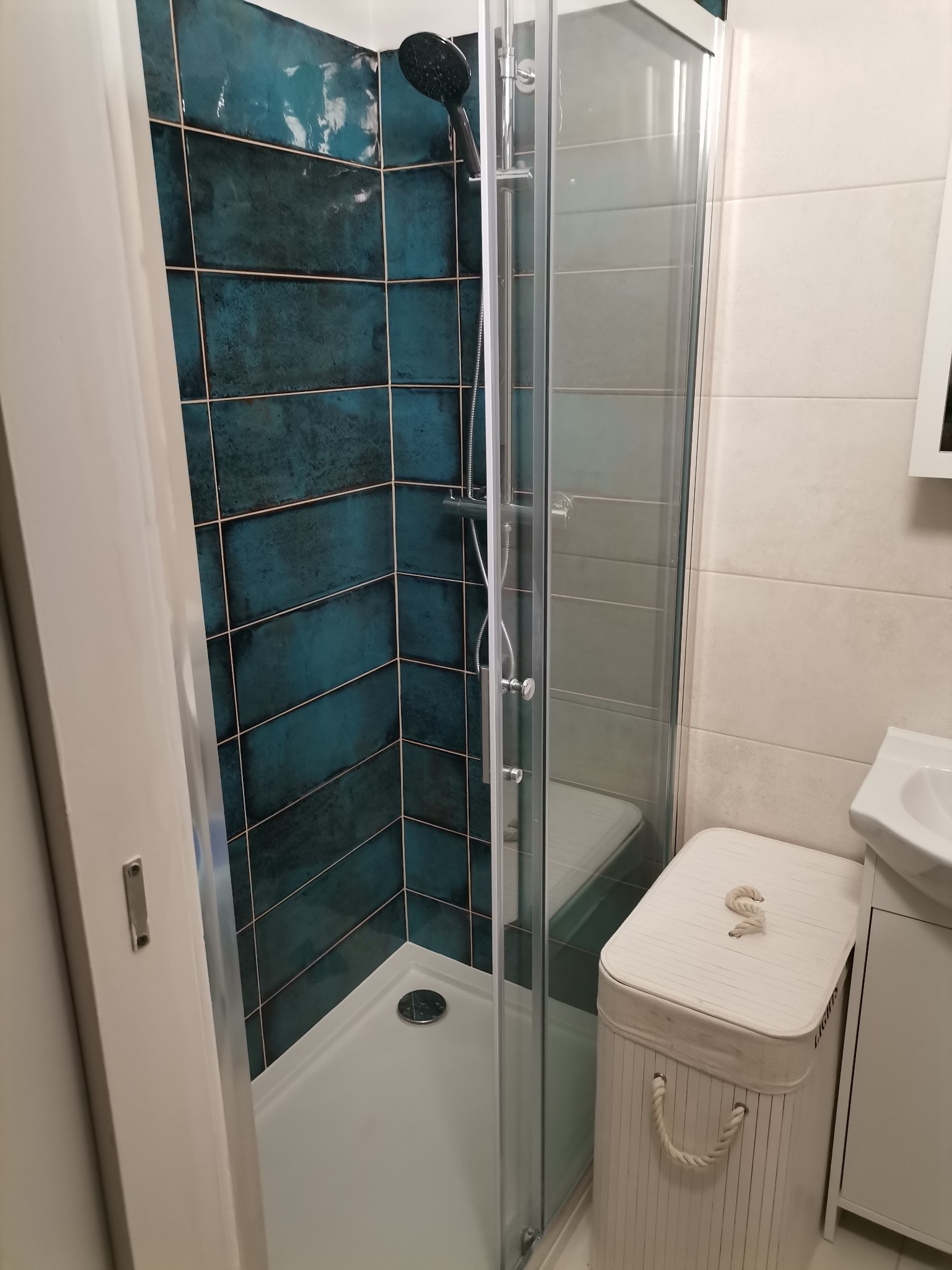 Kabina prysznicowa z turkusowymi, błyszczącymi płytkami ułożonymi w cegiełkę, obok biała umywalka i kosz na pranie z sznurkowymi uchwytami.