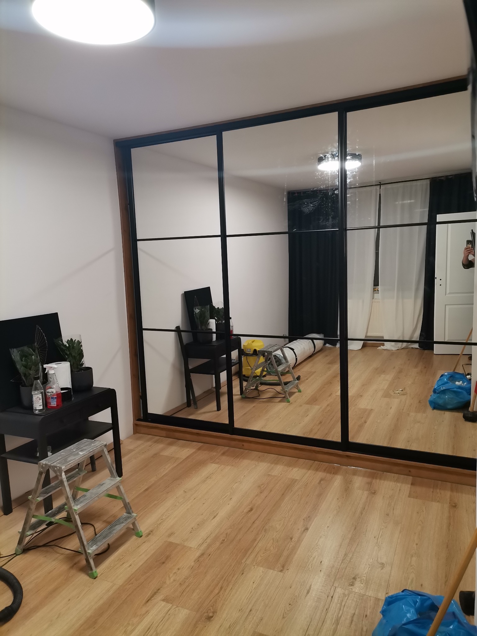 Montaż dużej szafy przesuwnej z lustrami w czarnej ramie, widoczne narzędzia i akcesoria montażowe, podłoga wyłożona panelami.