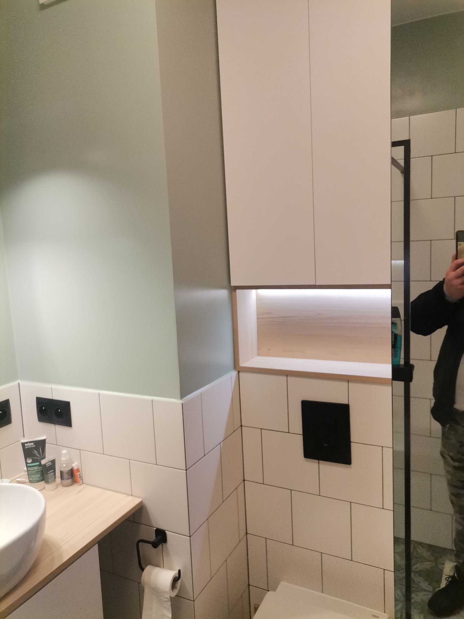 Nowoczesna łazienka z białymi, prostokątnymi płytkami, podwieszaną szafką z oświetleniem LED, umywalką nablatową i toaletą podwieszaną z czarnym przyciskiem spłukującym.