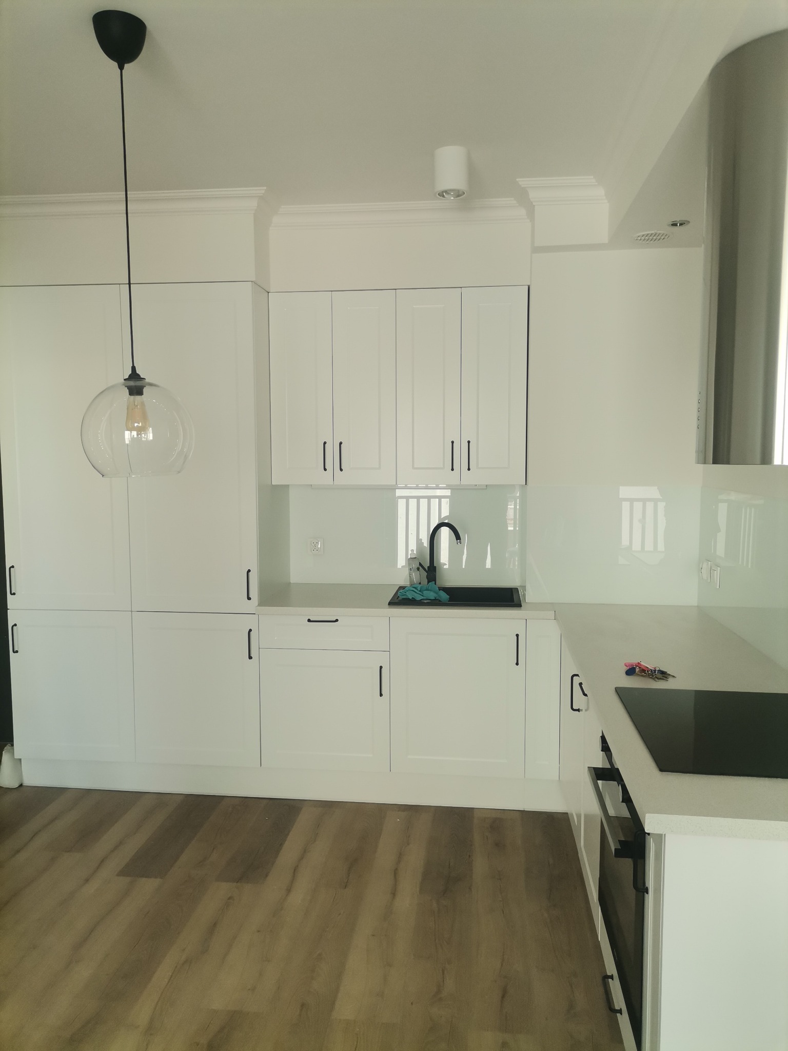 Nowoczesna, biała kuchnia z drewnianą podłogą, czarnymi uchwytami szafek i szklanym panelem nad blatem, oświetlona wiszącą lampą z przezroczystym kloszem