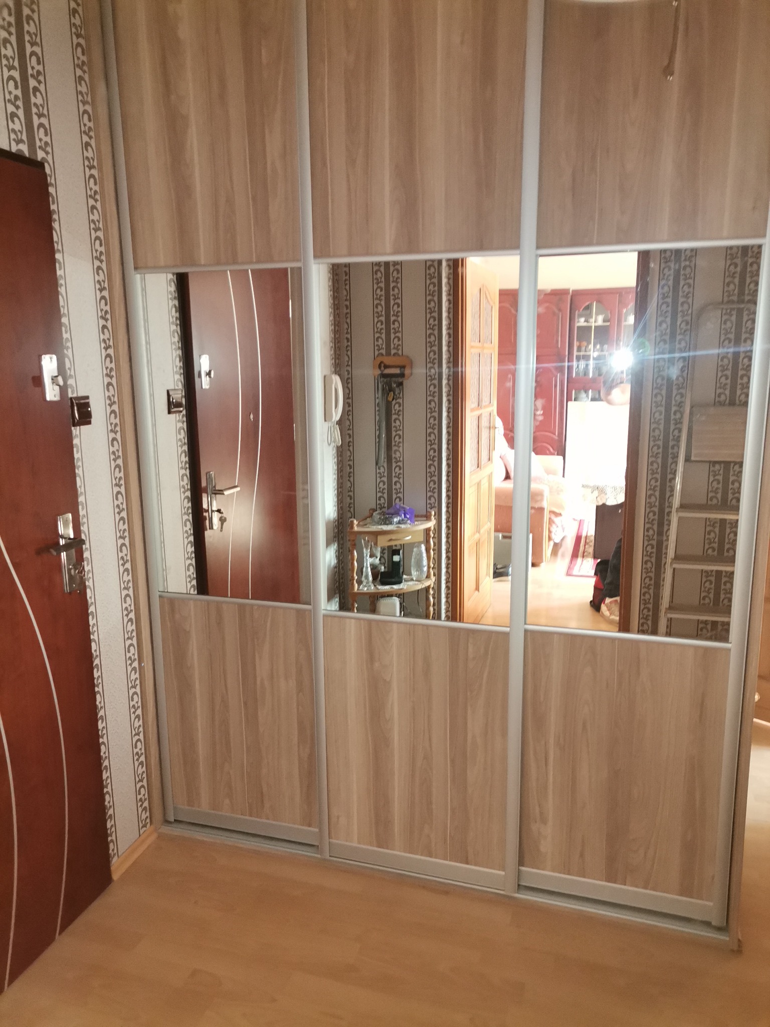 Duża szafa przesuwna z lustrami w przedpokoju, odbicie wnętrza mieszkania widoczne w lustrach, brązowe drzwi wejściowe po lewej stronie.