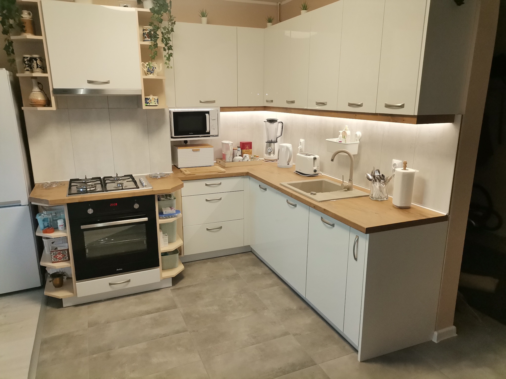 Jasna kuchnia z białymi szafkami i drewnianymi blatami, oświetlenie LED pod szafkami wiszącymi, widoczny piekarnik Amica, kuchenka gazowa, zlew, mikrofala i akcesoria kuchenne.